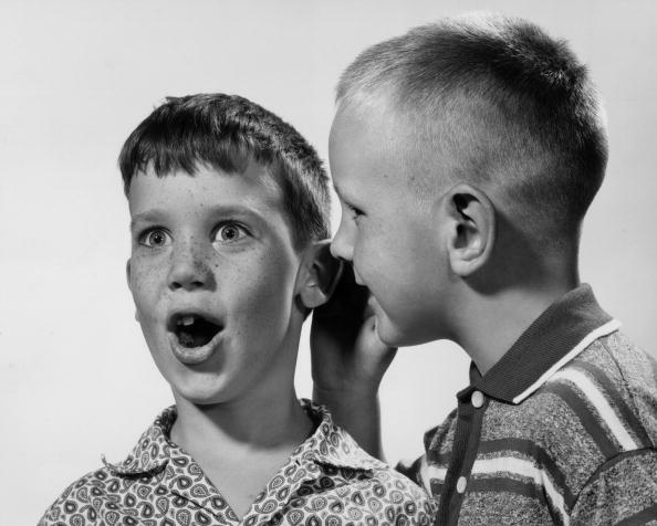 circa 1955:  Studio headshot of one young boy whispering a secret in another boy's ear, who looks surprised. There is a white