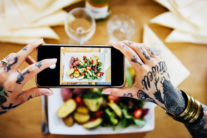 Overhead shot of tattooed hands of a young woman taking pictures of her dinner plate