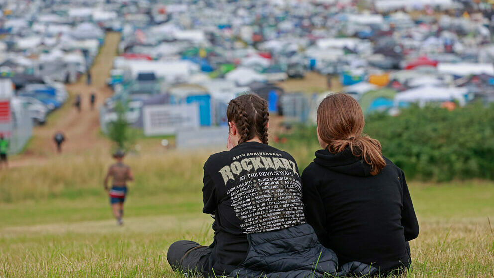 Zwei Festival-Besuchenden sitzen gegenüber dem Zelt- und Campingplatzgelände auf dem Flugplatz. Zum Festival Rockharz in Ba